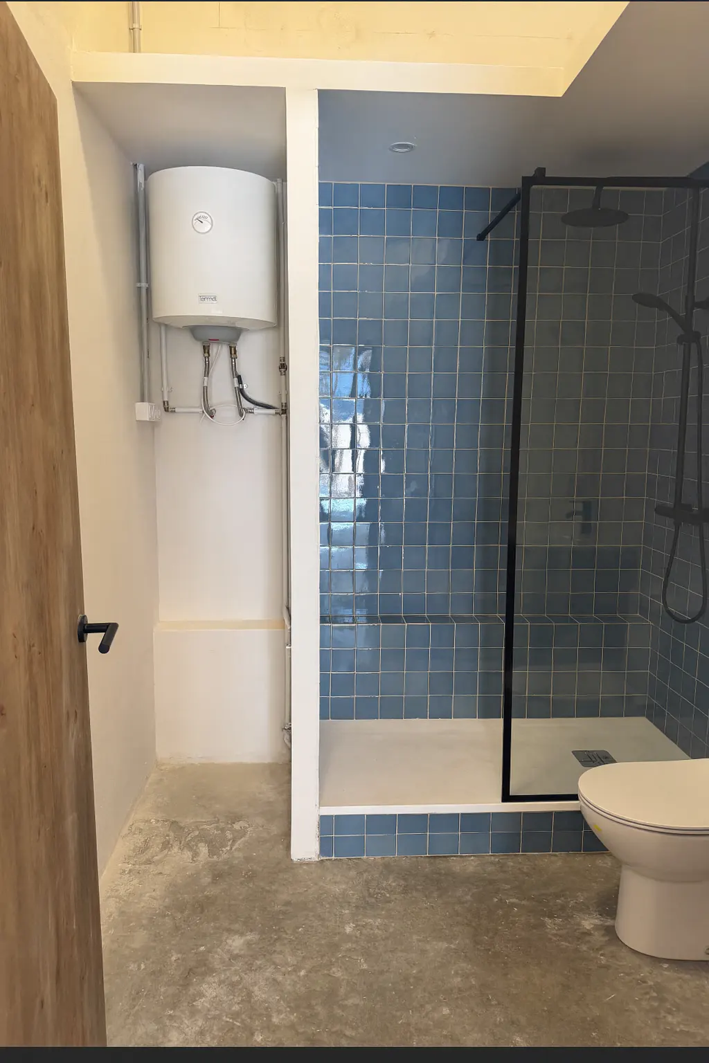 Bathroom with blue tiled shower, black frame, and white toilet. A water heater is in the adjacent space.