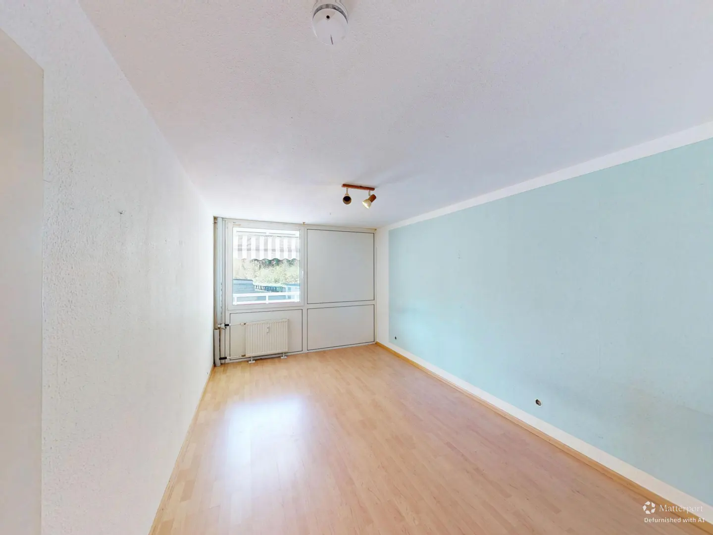 A bright, empty room with light wood floors, white walls, and a light blue accent wall. A window with a striped awning is visible.