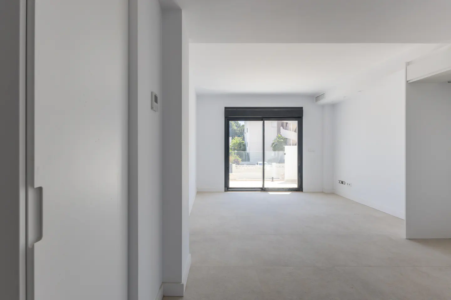 Empty, minimalist room with white walls, light tile floors, and a large sliding glass door leading to an outdoor area.