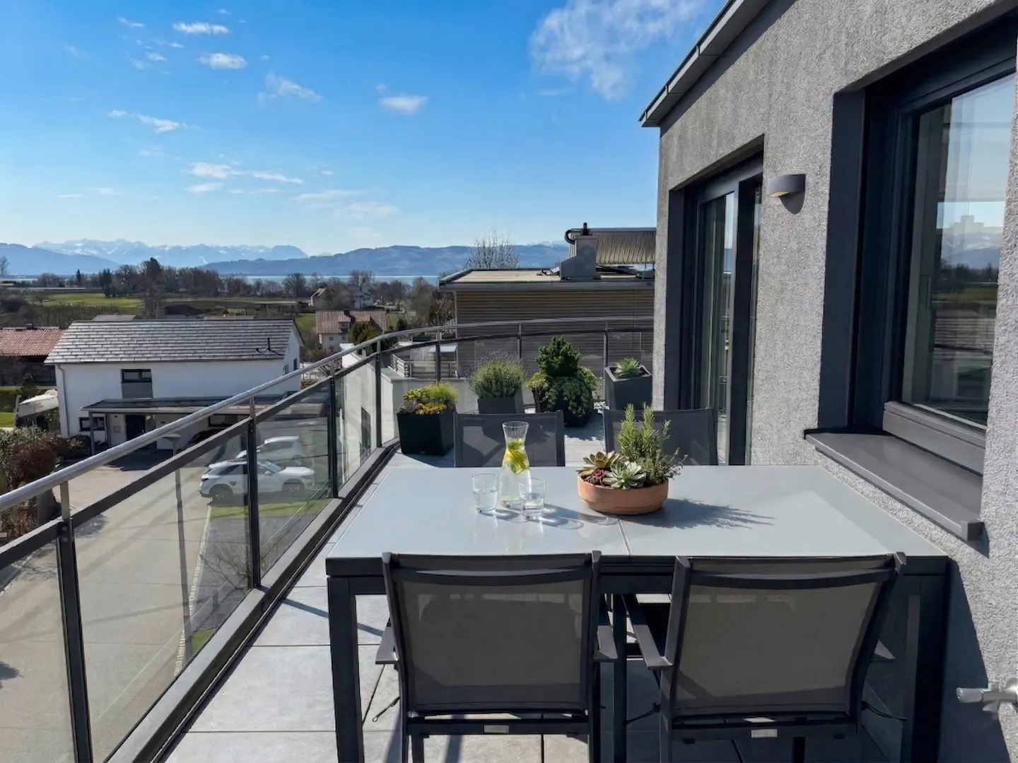 Rooftop patio with a gray table, chairs, and plants overlooking a scenic view of houses, fields, and distant mountains under a blue sky.