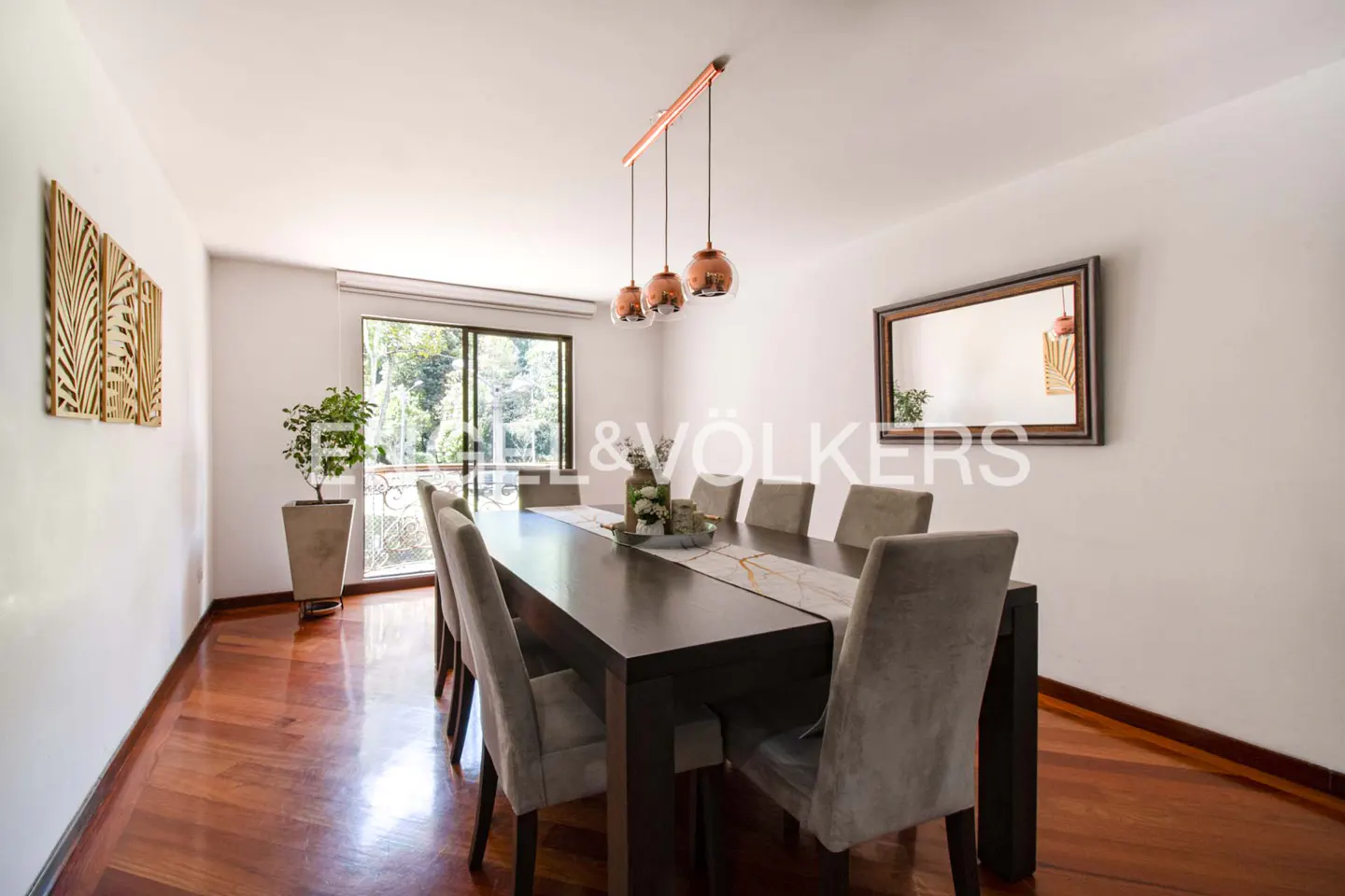 Dining room with a long dark wood table, eight gray chairs, and a white runner. Three copper pendant lights hang above. Hardwood floors and white walls.