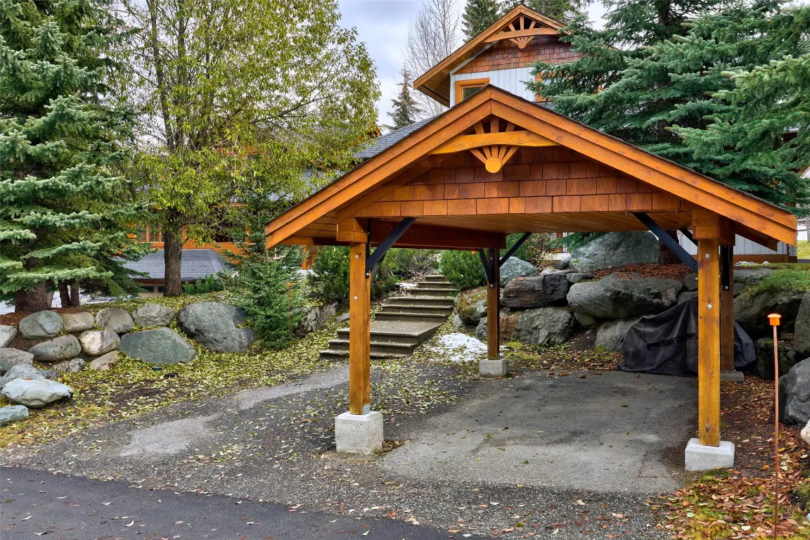A covered parking area with a wood roof and support beams, surrounded by trees and rocks. Steps lead up to a house in the background.