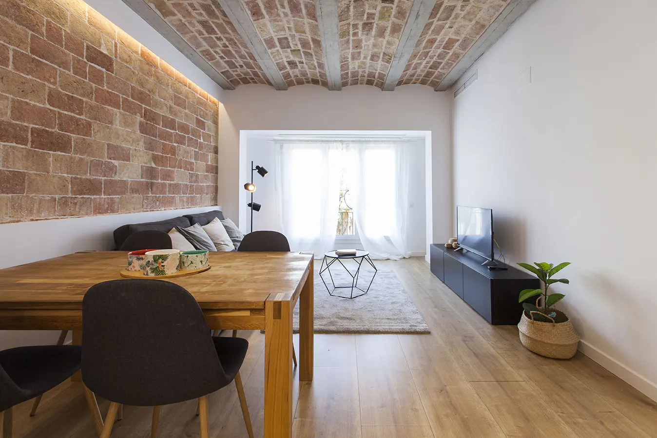 Bright living room with brick wall, wood floors, and a table with chairs. A sofa and TV are also visible.