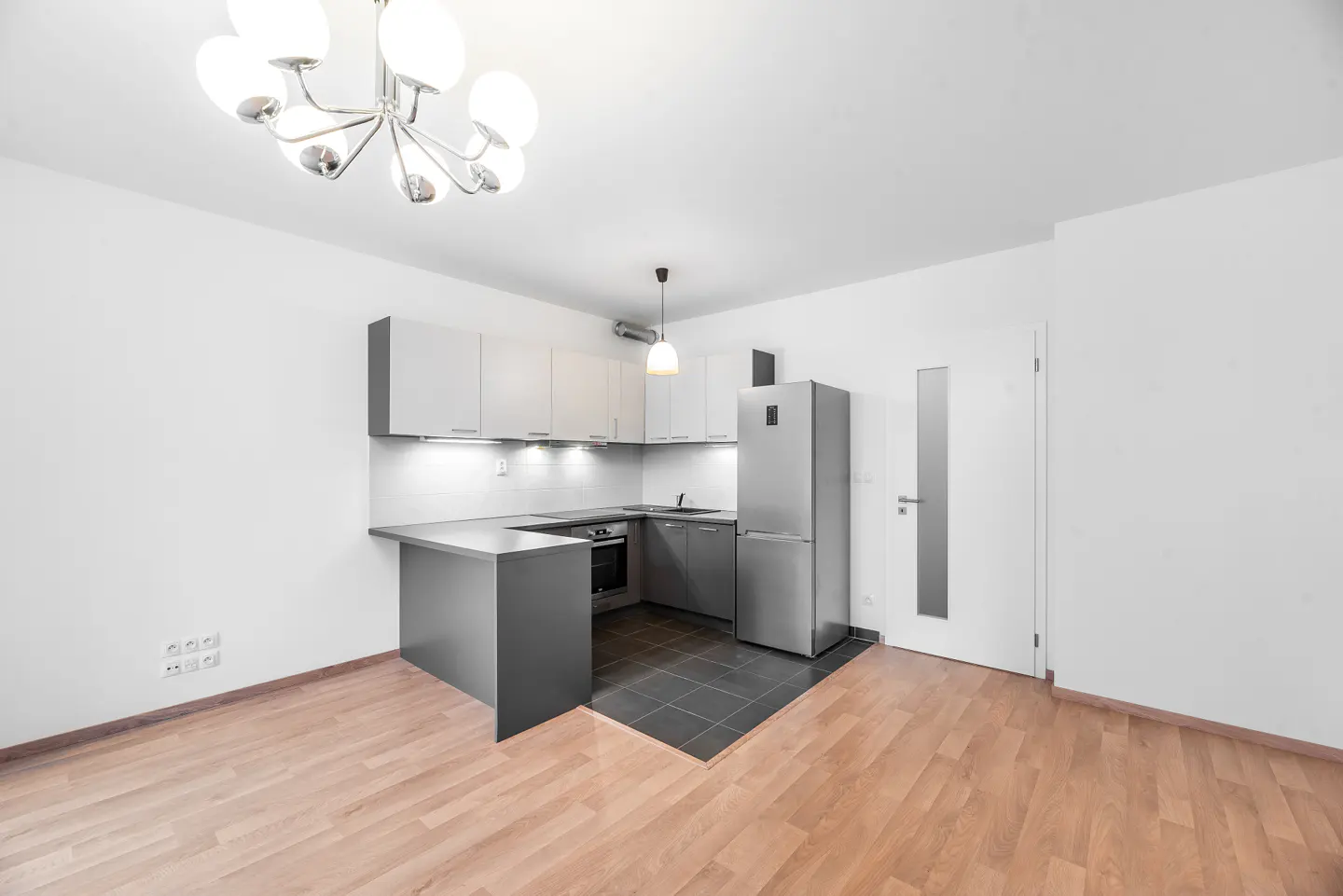 Modern kitchen with white walls, grey cabinets, stainless steel fridge, and a chandelier over light wood flooring.
