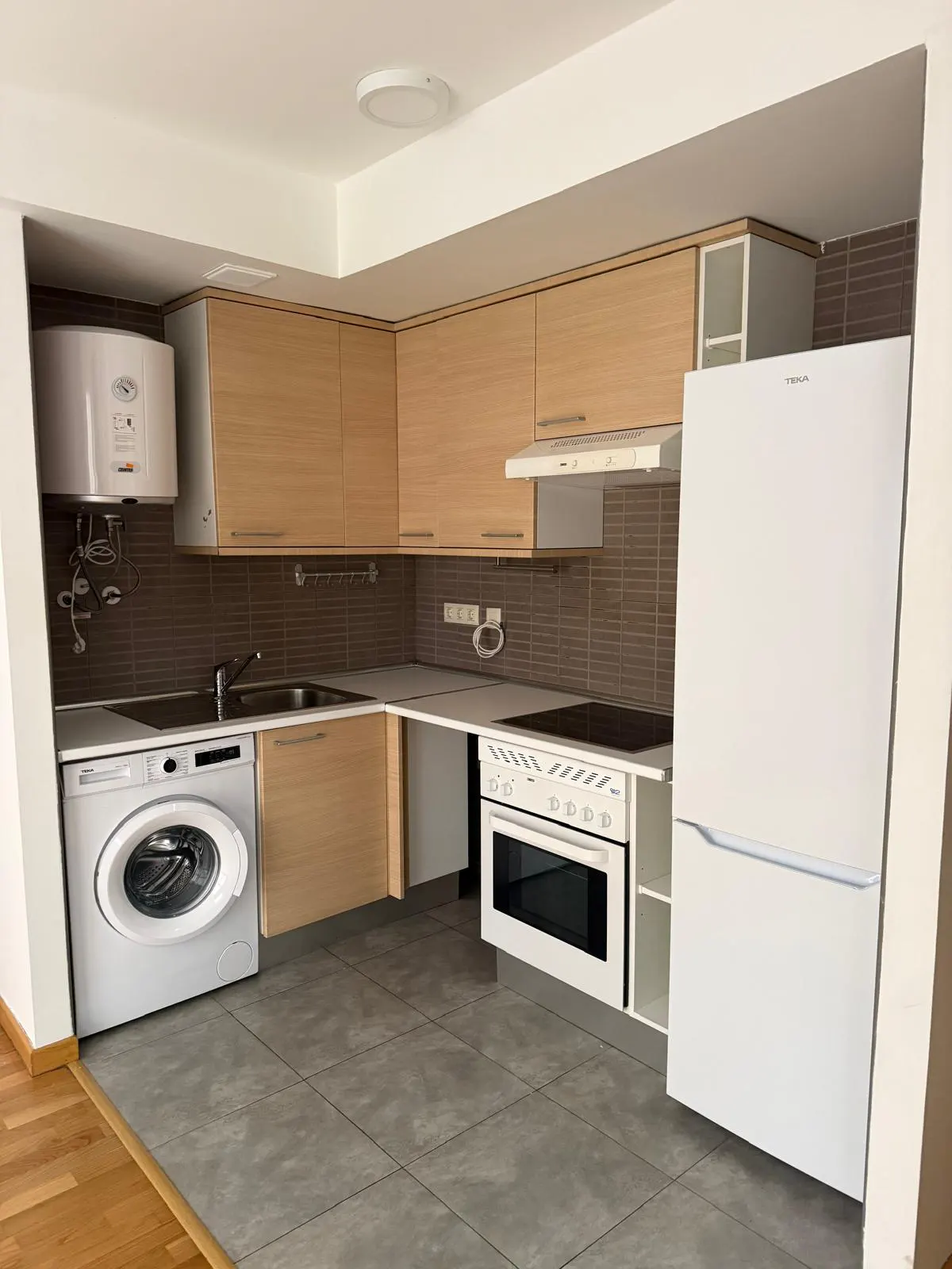 A modern kitchen with light wood cabinets, white appliances, and gray tile flooring. A washing machine is next to the sink.