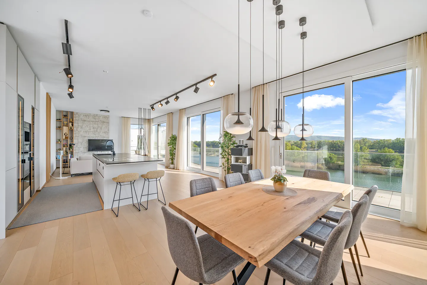 Bright, modern apartment with a wood dining table, gray chairs, and pendant lights. Large windows offer a view of a lake and trees.