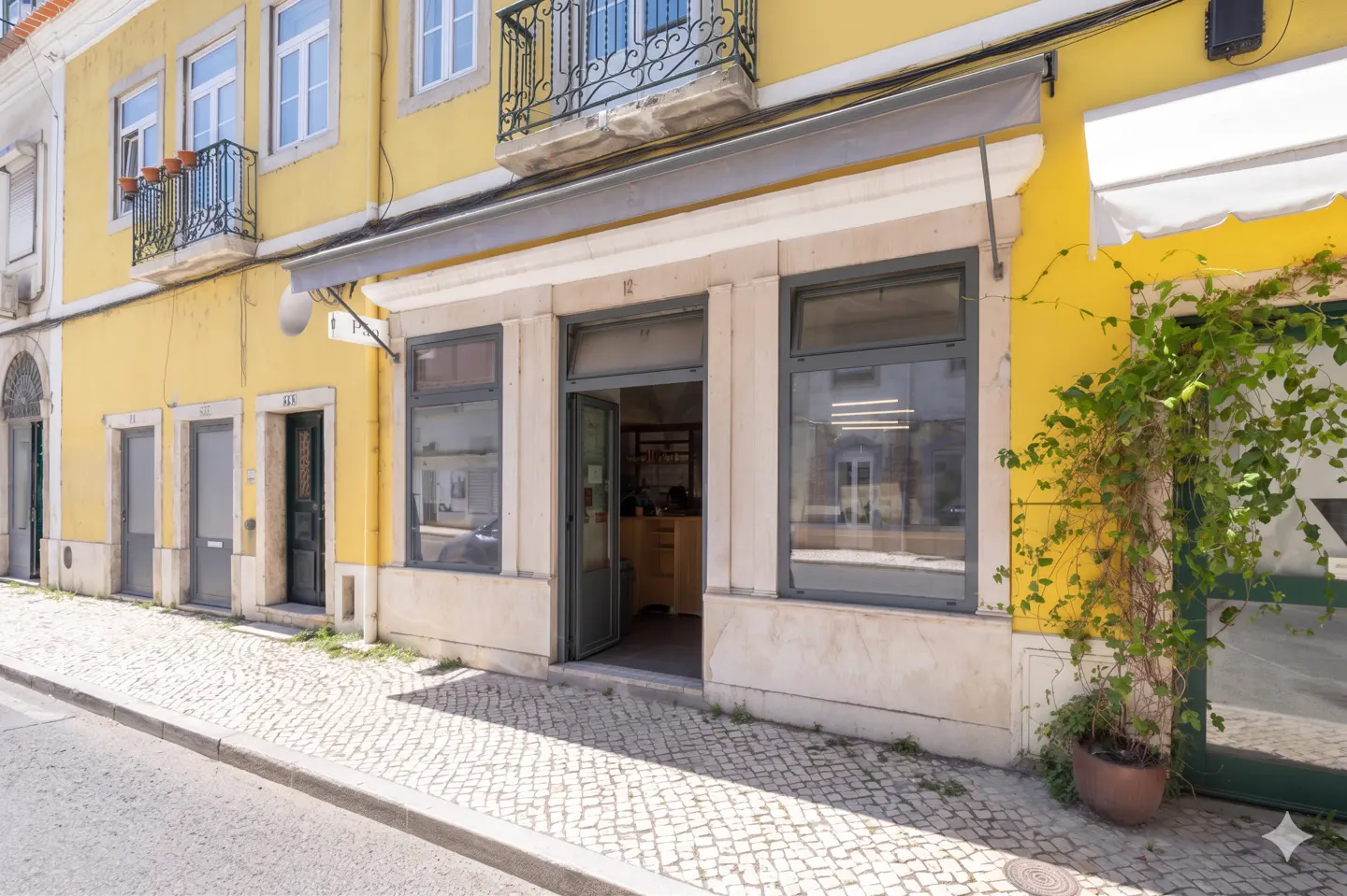 Exterior view of a yellow building with an open storefront. Cobblestone street in front. Balcony with black railing above.
