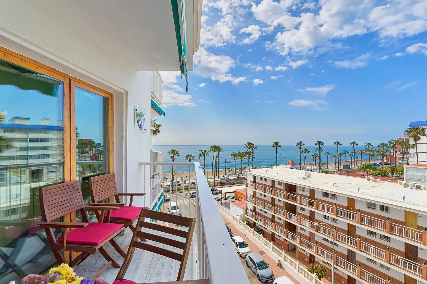 Balcony view with wooden chairs, red cushions, and flowers overlooking the ocean, palm trees, and buildings under a blue sky.