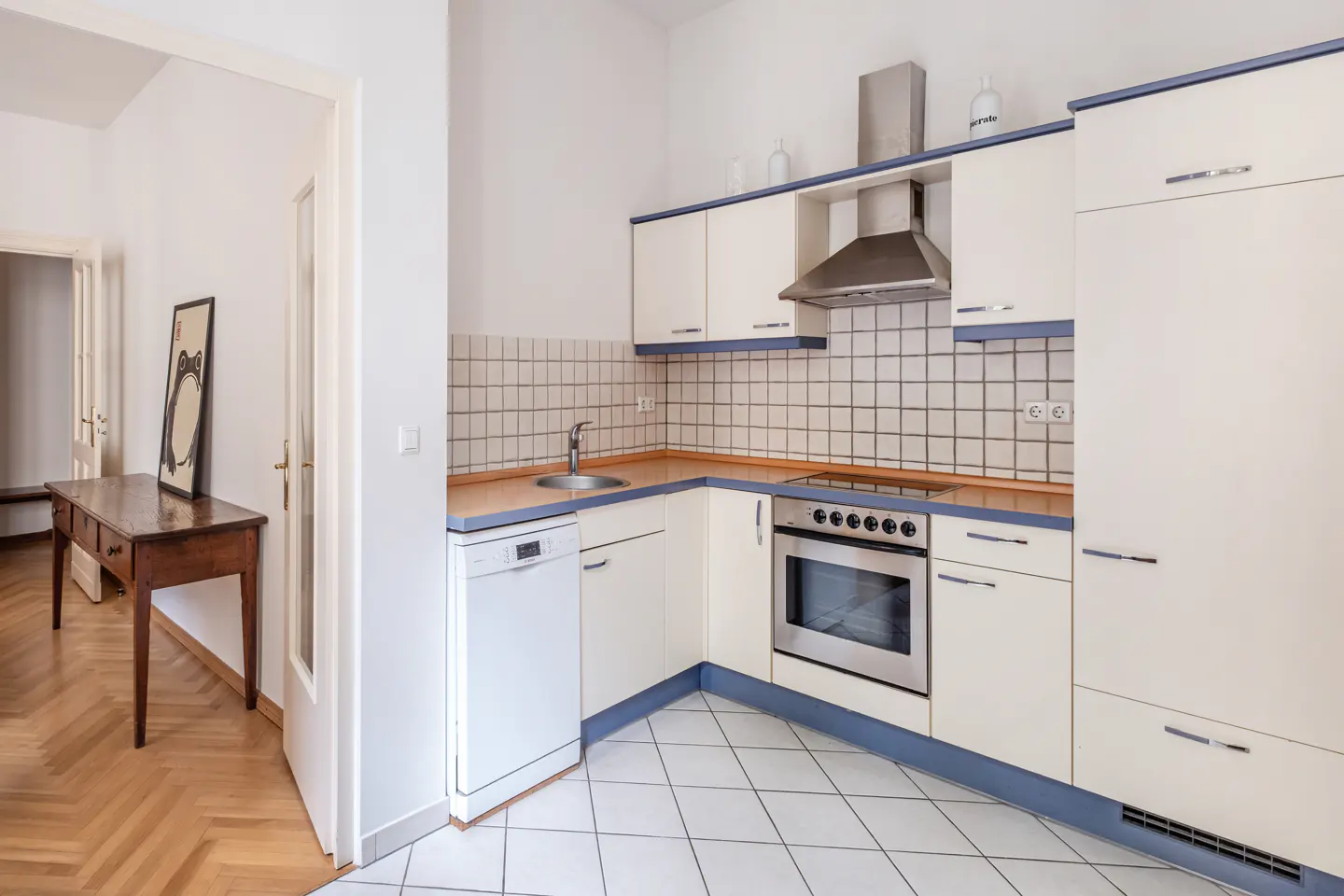 Modern kitchen with cream cabinets, blue trim, stainless steel appliances, and white tiled floors, next to a hallway.