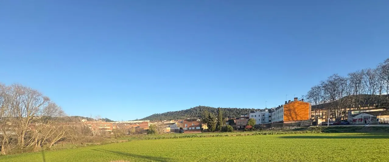 Landscape view of a green field, buildings, and a mountain under a clear blue sky.
