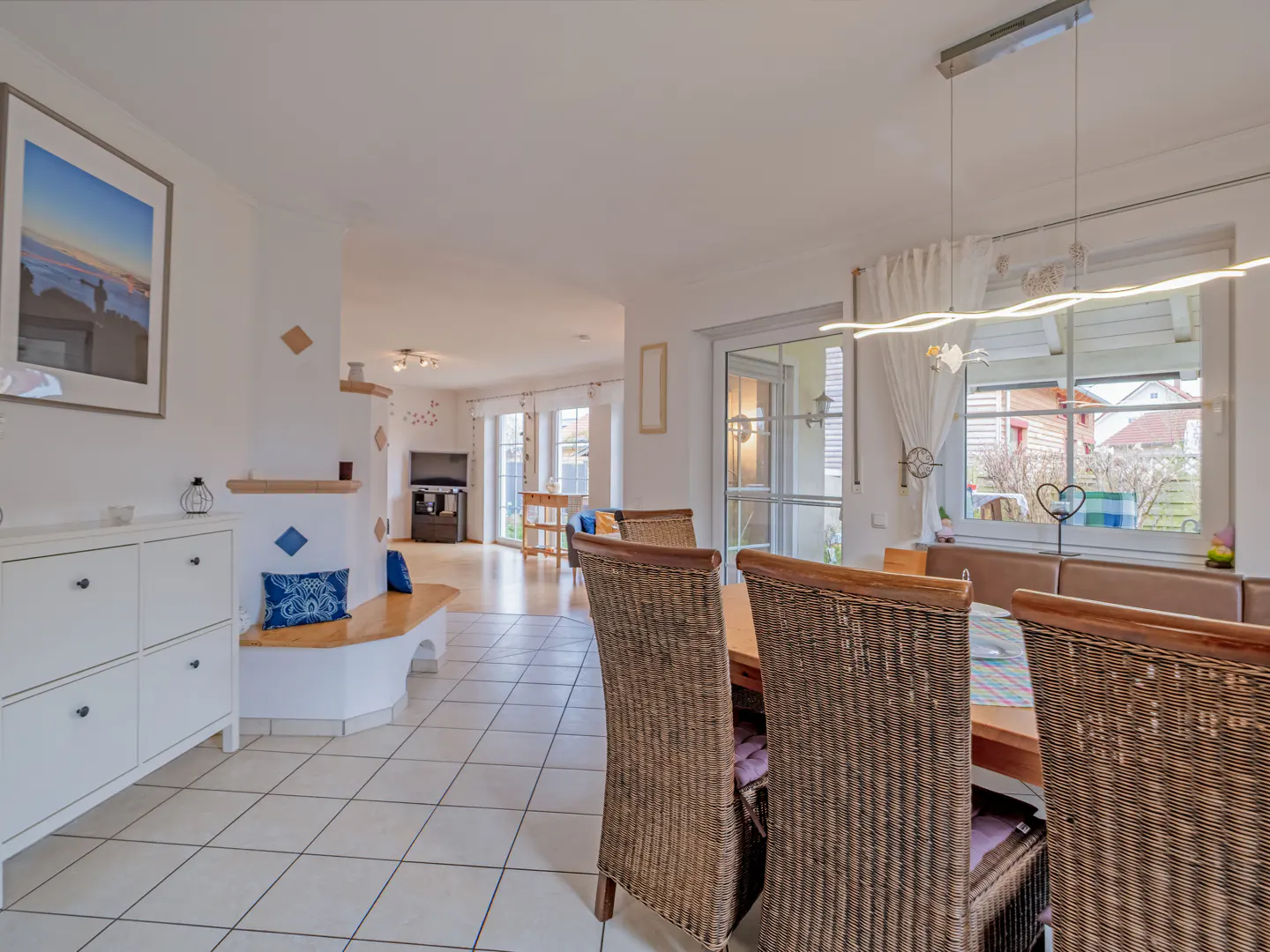 Bright, open-concept dining area with wicker chairs, a wooden table, and a bench seat by a large window. White walls and tile floors create a clean, airy feel.