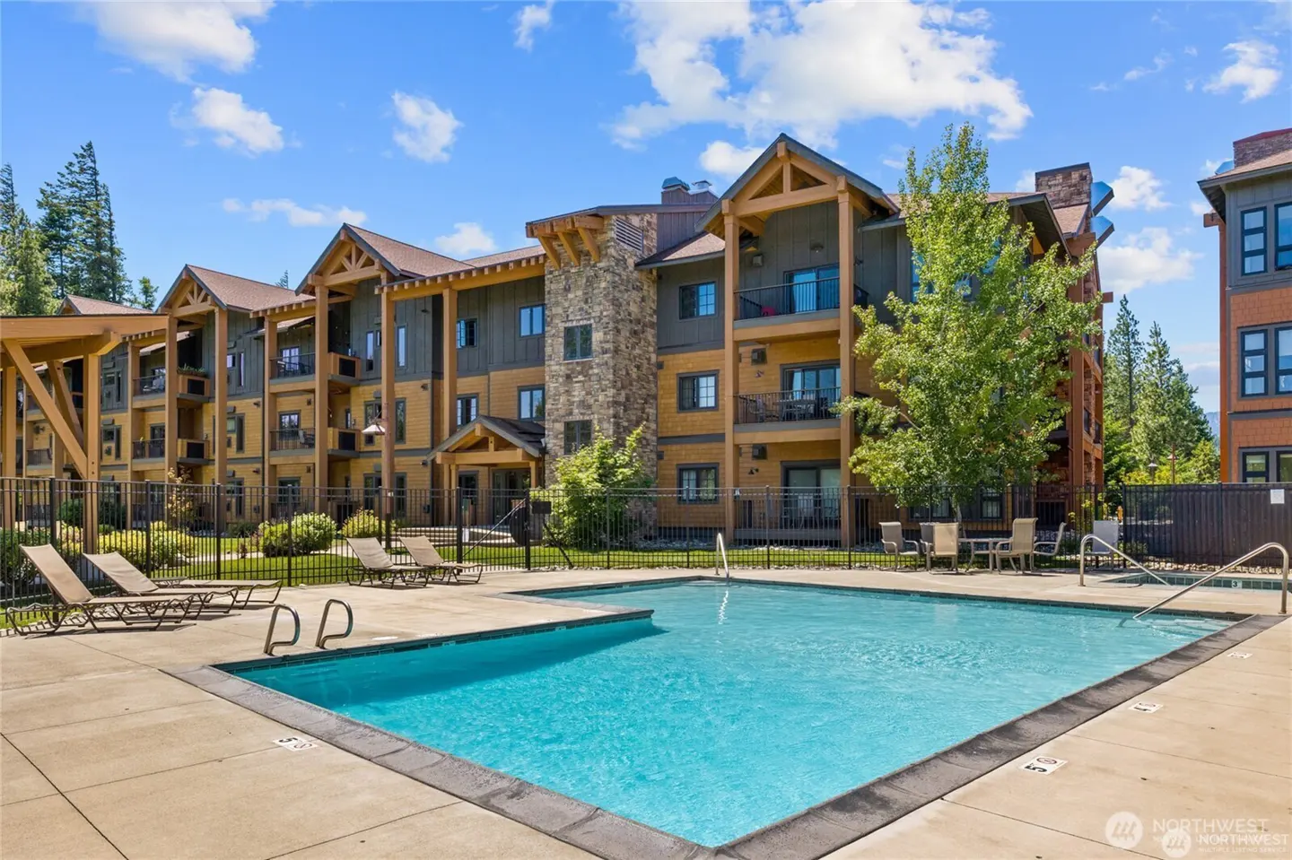 A multi-story apartment building with stone and wood siding overlooks a swimming pool with lounge chairs on a sunny day.