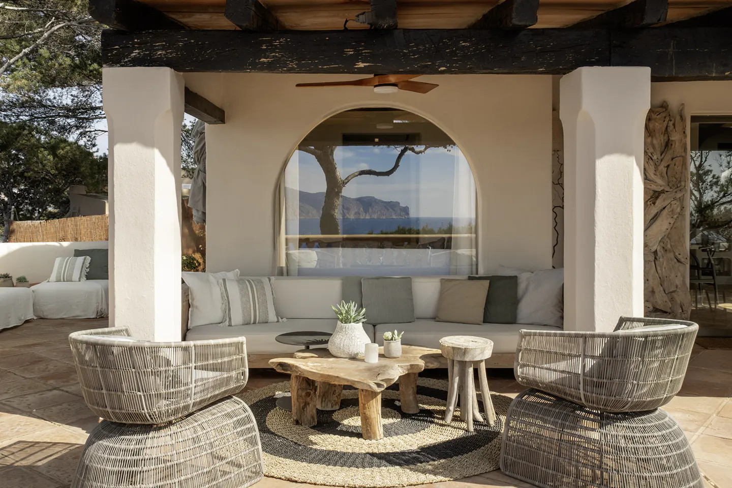 Outdoor patio with white stucco columns, a wooden pergola, and wicker furniture. A large arched window overlooks the ocean.