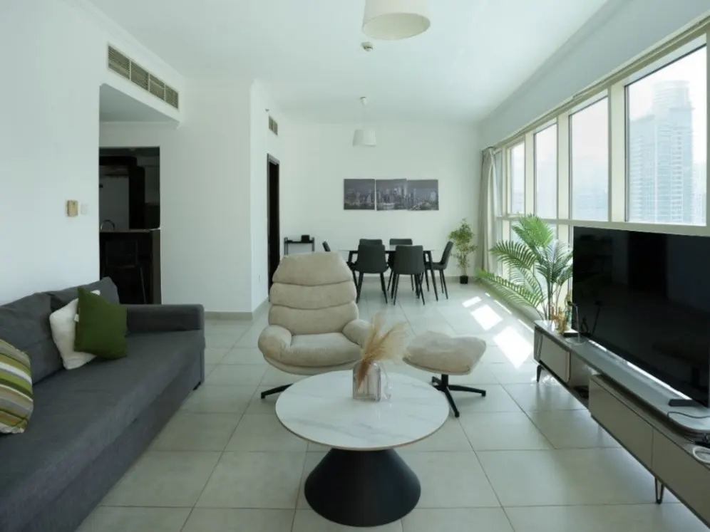 Bright, modern living room with gray sofa, beige chair, and dining table. Large windows offer city views. A TV and plants add to the space.