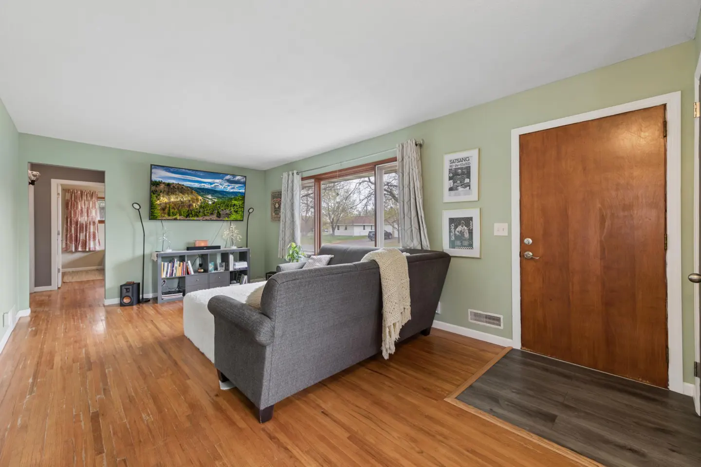 Living room with hardwood floors, gray sofa, TV showing a mountain scene, and light green walls. A wooden door is on the right.