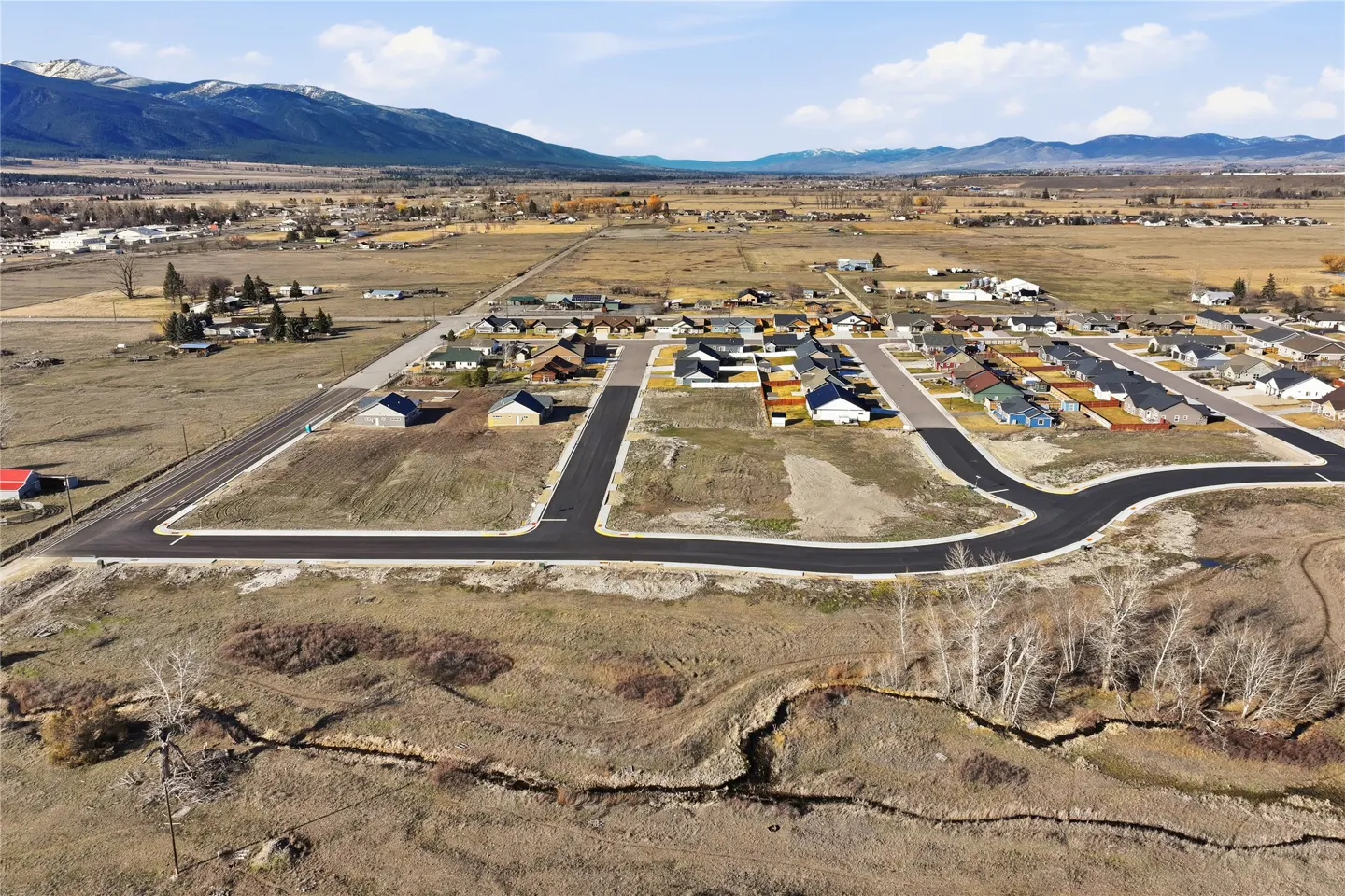 Aerial view of a residential area with houses, roads, and vacant lots, set against a backdrop of mountains and a blue sky.