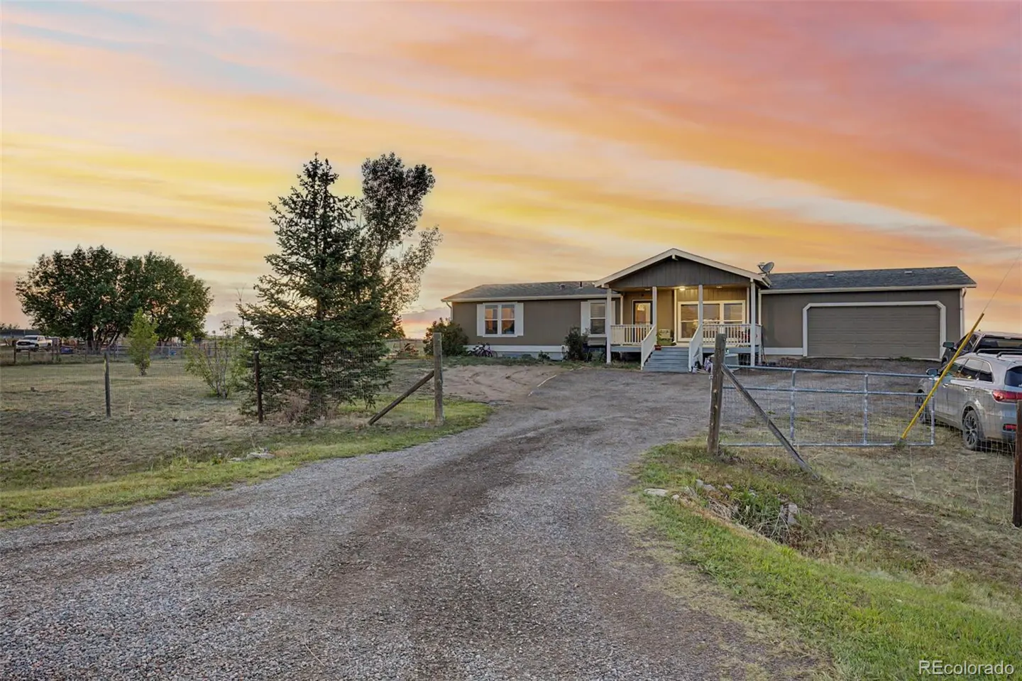 Single-story ranch home with a gravel driveway and attached garage under a vibrant sunset sky in a rural setting.
