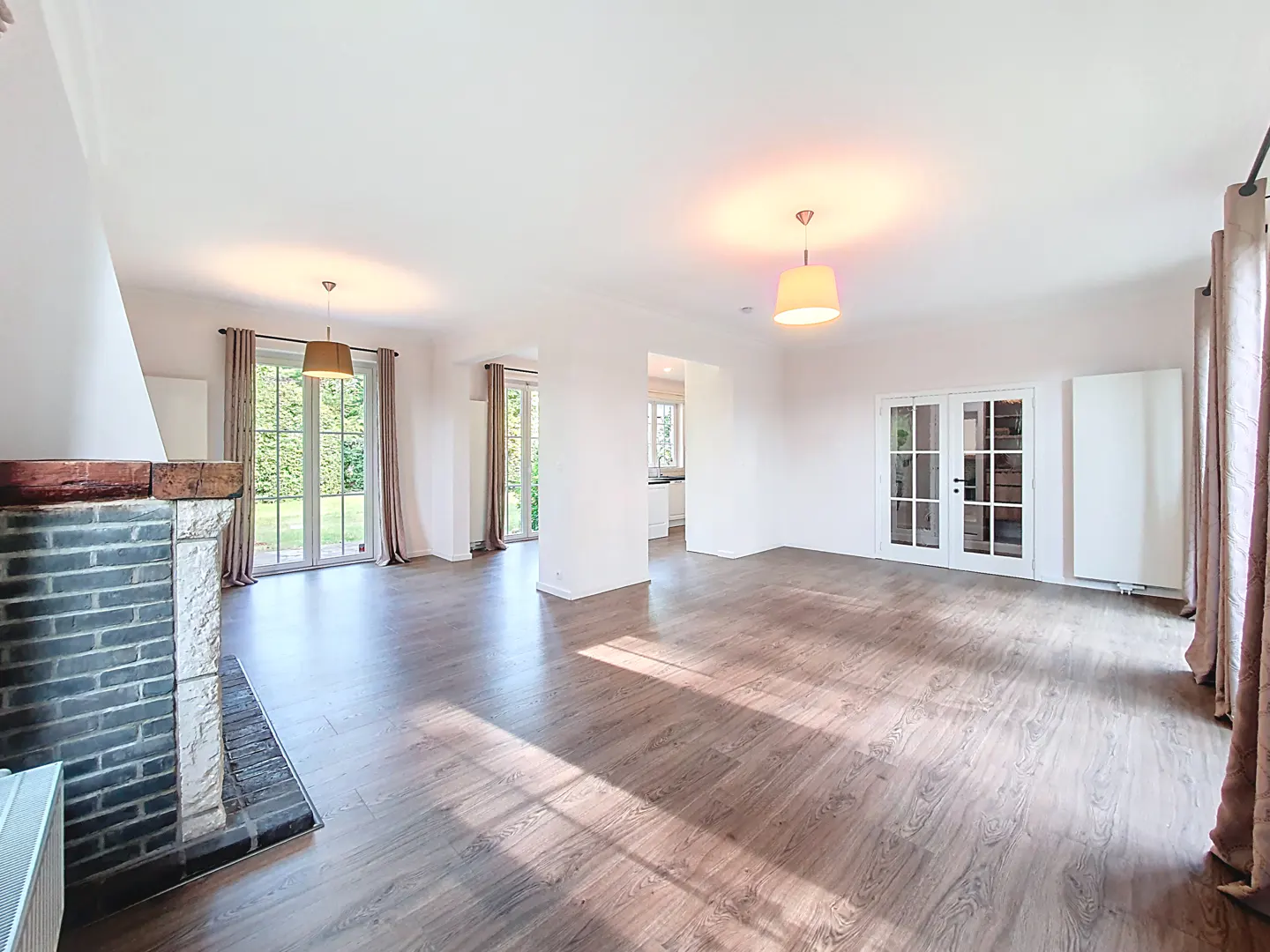 Bright, empty living room with wood floors, brick fireplace, and French doors to a green yard.