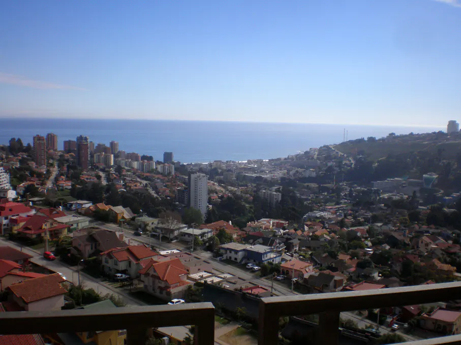 View of a coastal city with red-roofed houses, tall buildings, and the blue ocean under a clear sky.