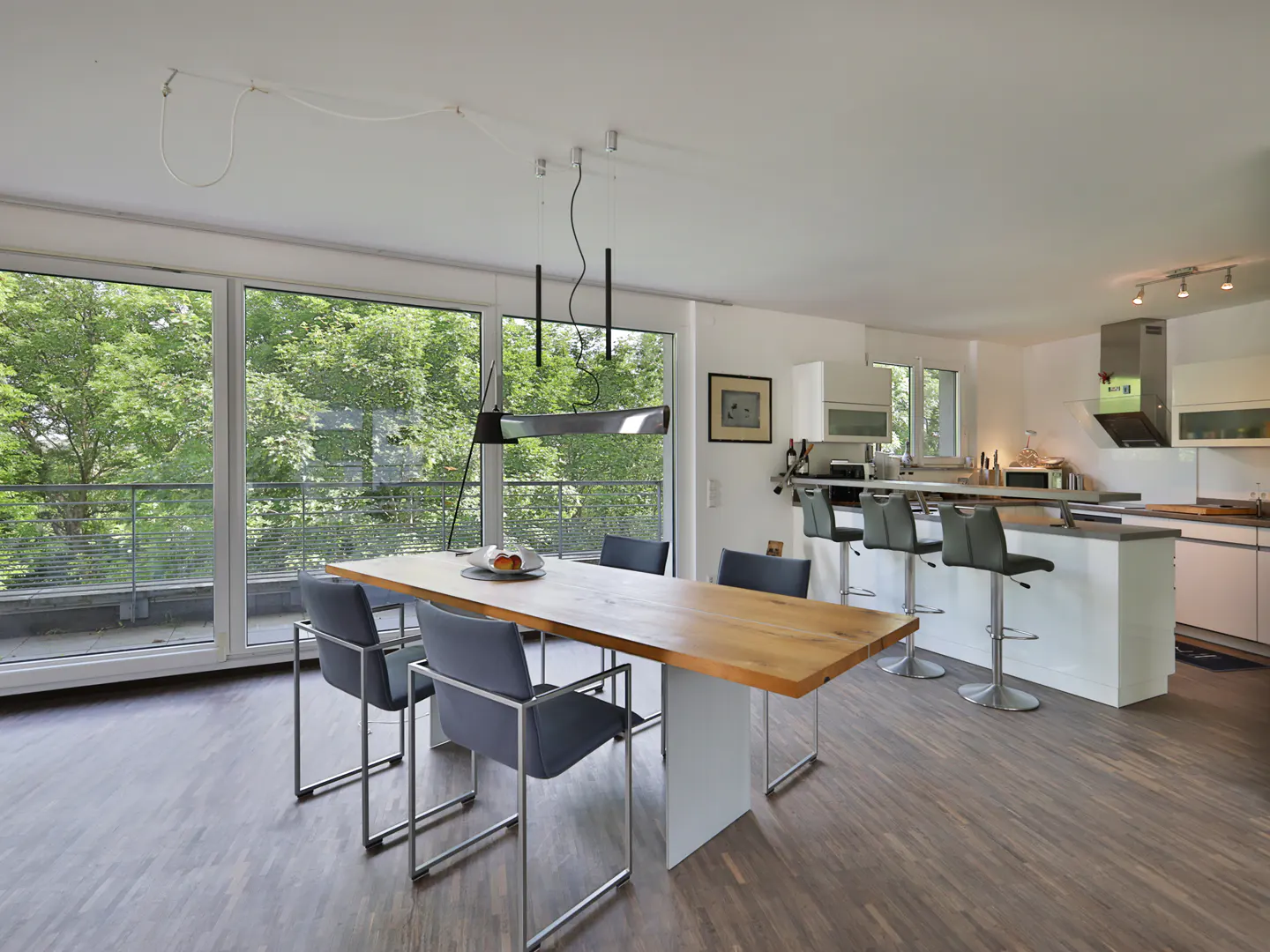Bright, open-concept dining and kitchen area with wood floors. A long wood table with gray chairs sits near large windows overlooking greenery. A kitchen island with bar stools is visible.