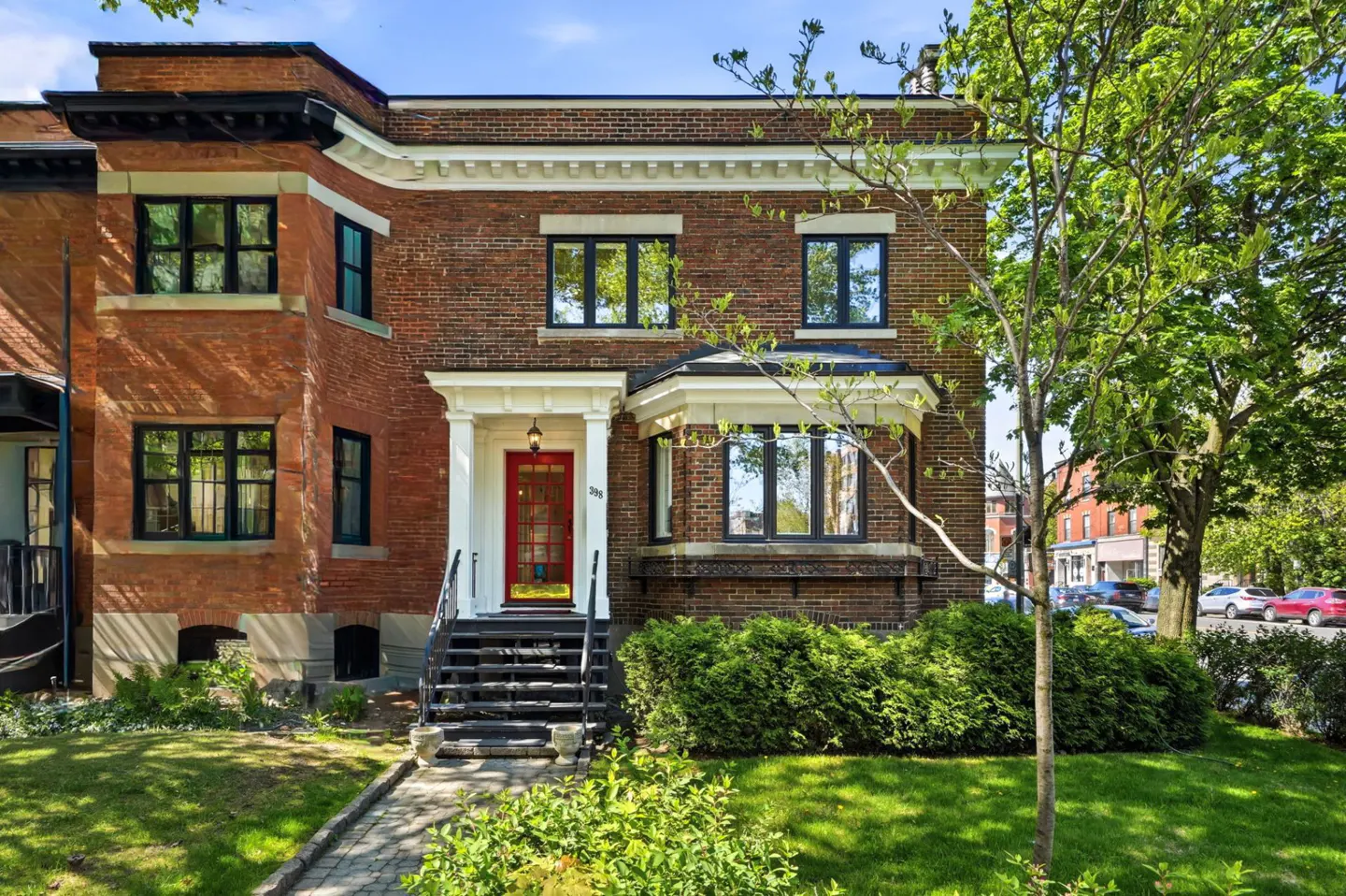 Two-story brick house with black windows and a red front door. A stone walkway leads to the front steps. Green lawn and trees.