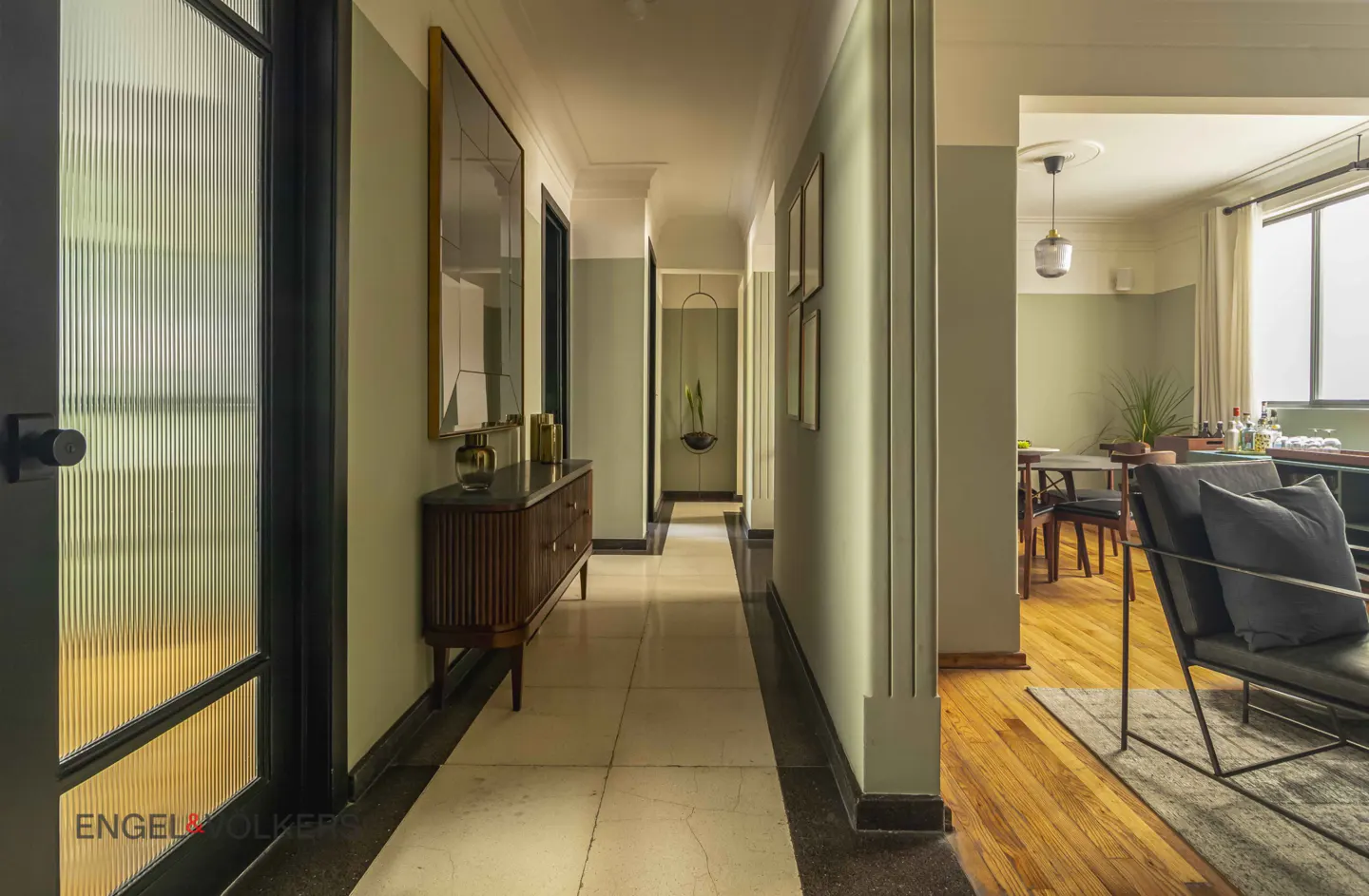 Hallway view with light green walls, a dark wood cabinet, and a textured glass door on the left. A dining area is visible at the end.