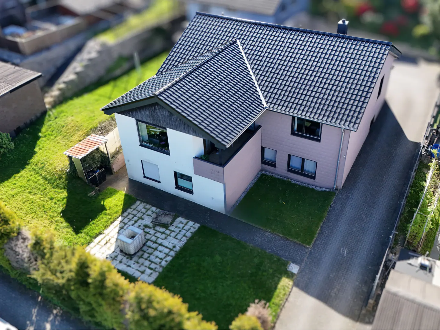 Aerial view of a two-story house with a dark gray roof, white and pink walls, green lawn, and a stone-paved patio.