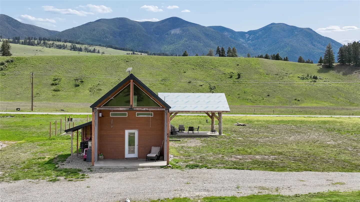 A small, brown house with a black roof sits in front of green hills and mountains. A covered patio is to the right of the house.