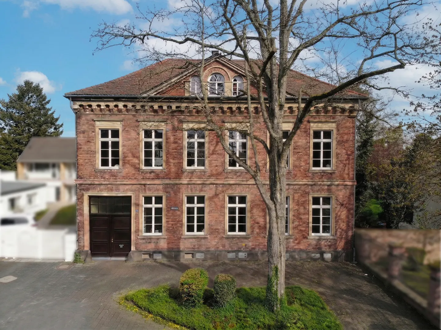 Two-story brick building with white-framed windows and a brown tile roof, partially obscured by a bare tree.