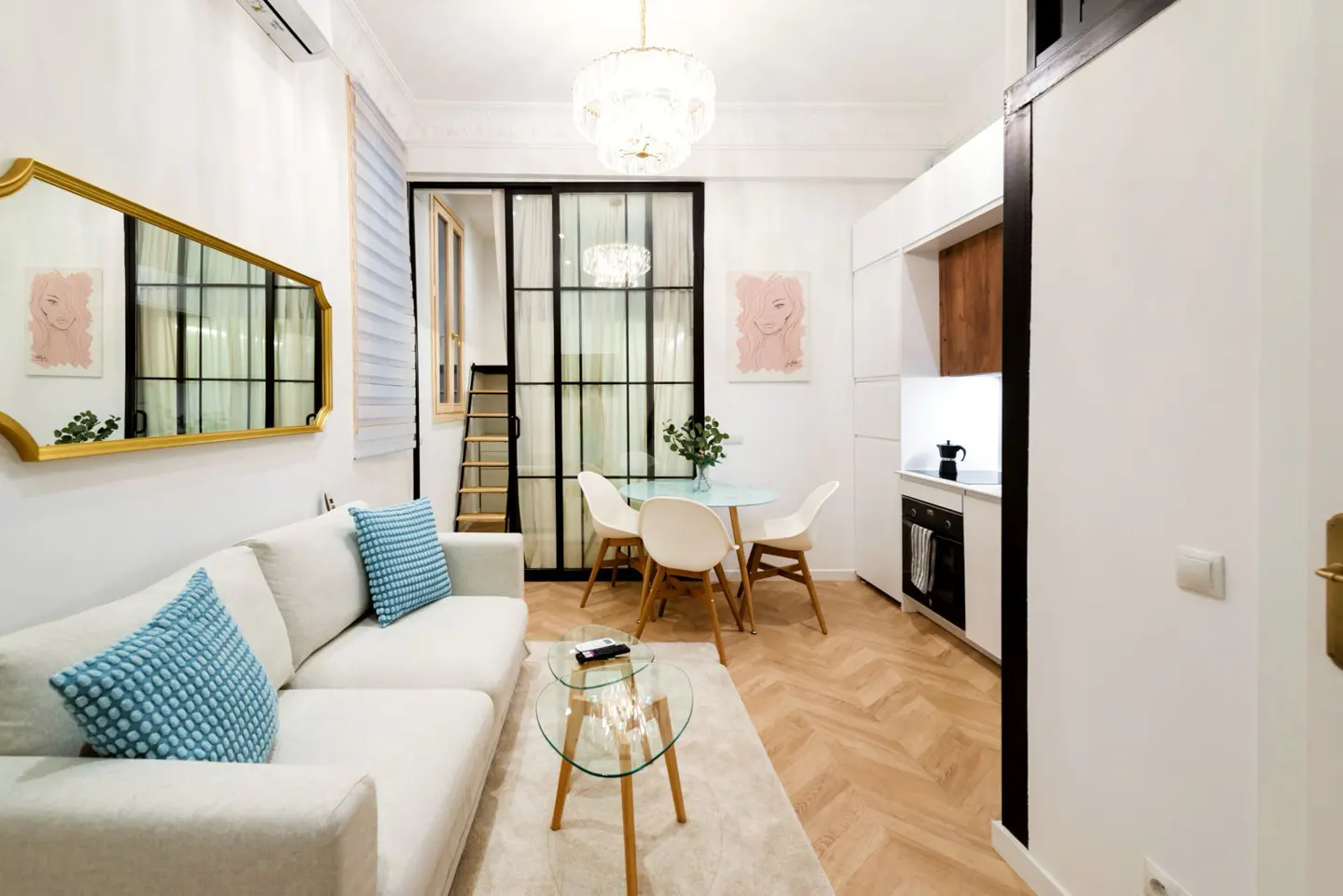 Bright apartment interior with white sofa, blue pillows, glass table, dining area, and compact kitchen. Herringbone wood floors and gold mirror.