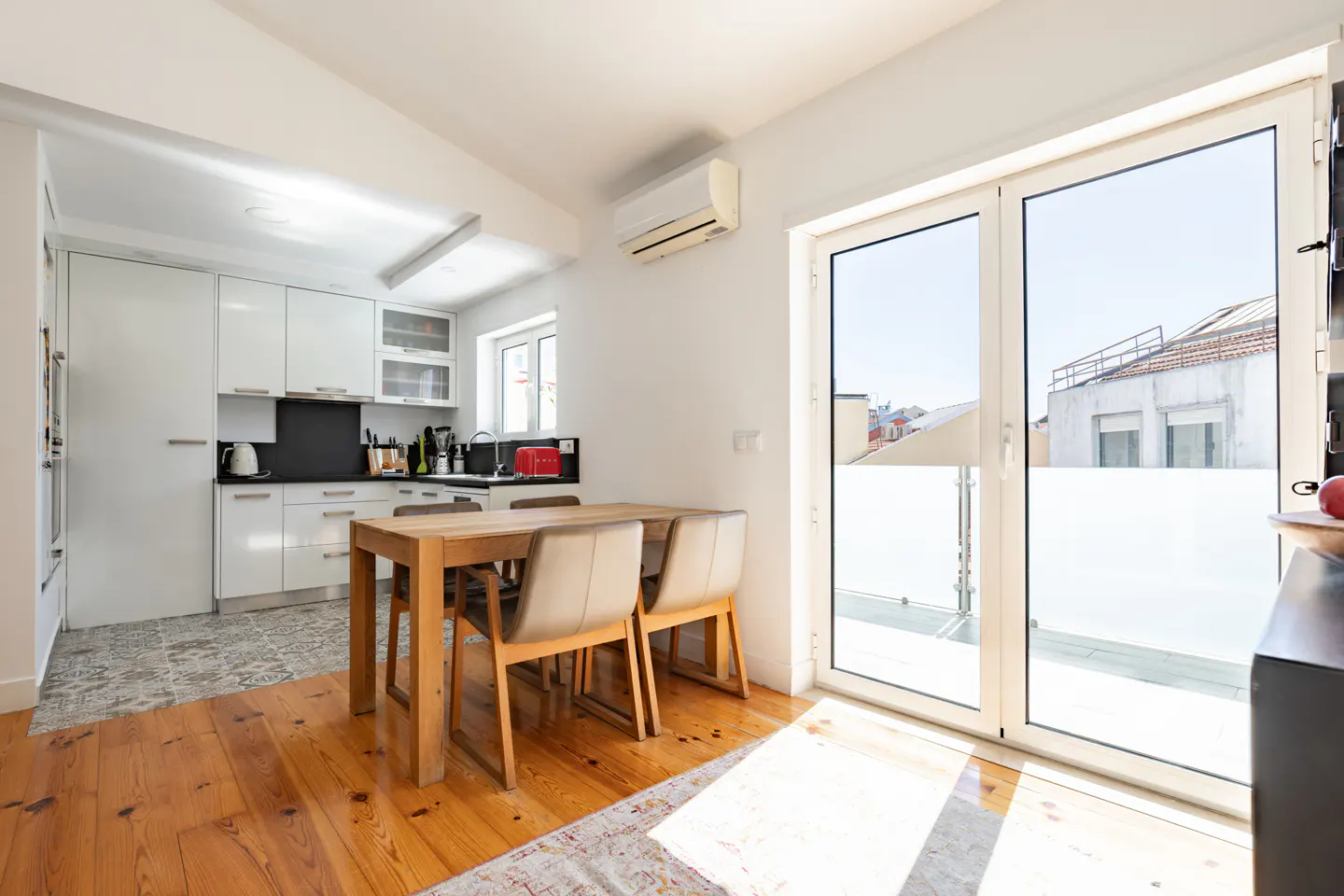 Modern kitchen with white cabinets, wooden dining table, and glass doors opening to a balcony with city views.