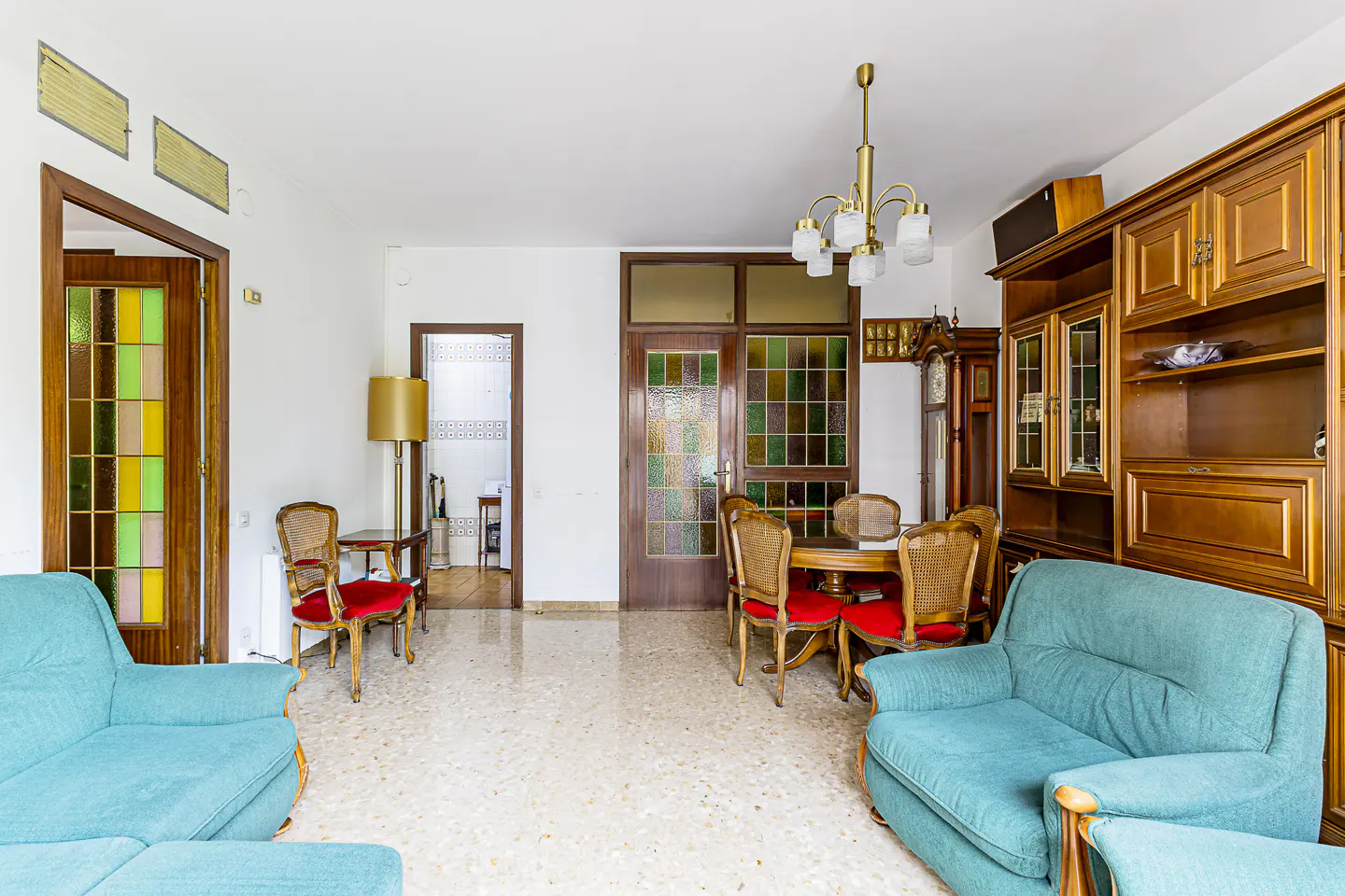 A bright living room with turquoise sofas, a wooden dining set, and a large wooden cabinet. A stained glass door adds color.