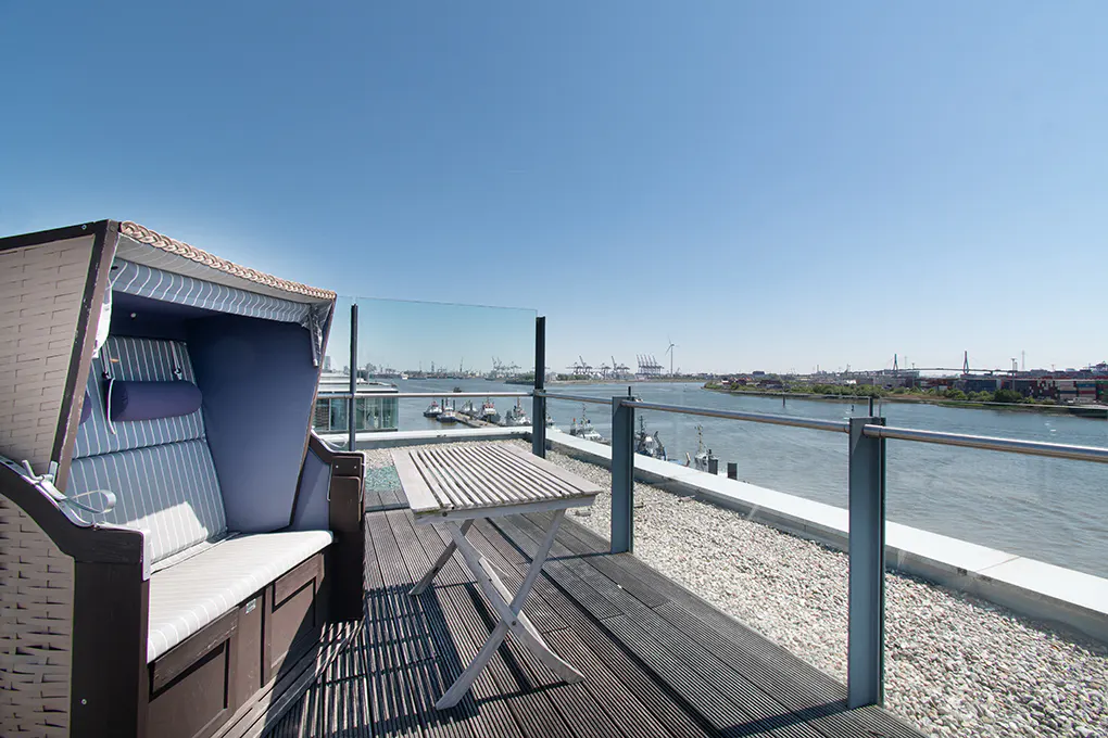 Rooftop patio with a wicker beach chair, small white table, and a view of a river and industrial port under a clear blue sky.