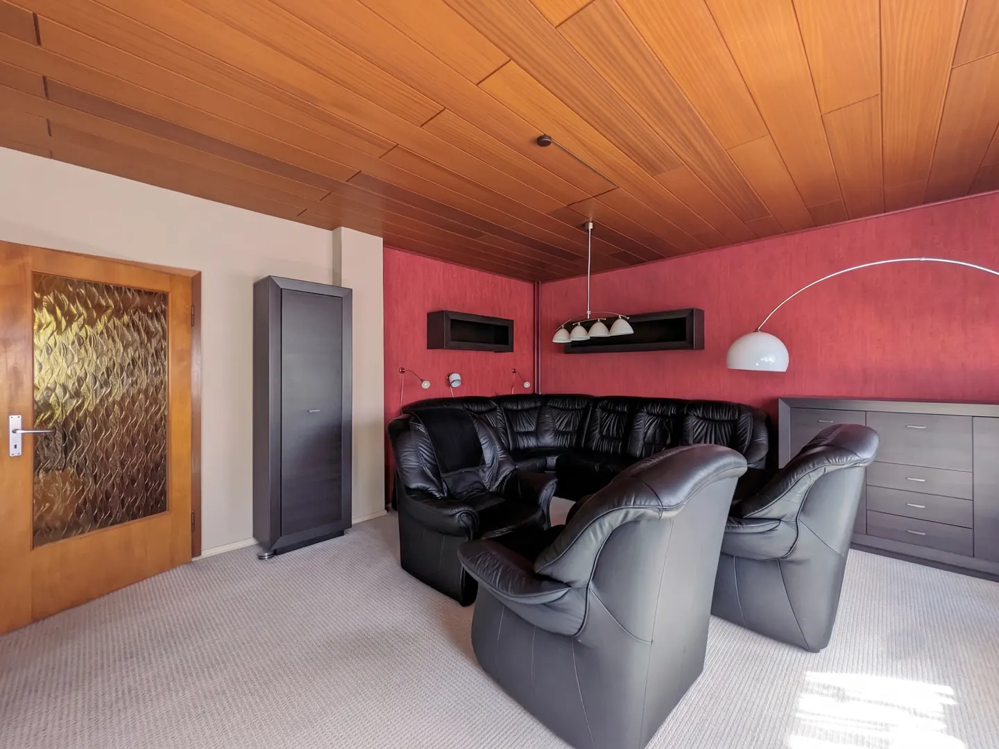 Living room with black leather sectional and armchairs, red accent wall, wood-paneled ceiling, and light gray carpet.