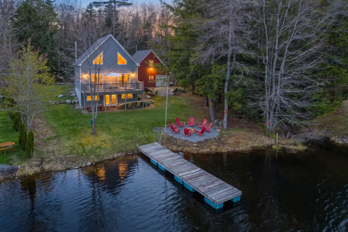 Lakefront home with a gray A-frame house, red garage, wooden dock, and red Adirondack chairs around a fire pit.