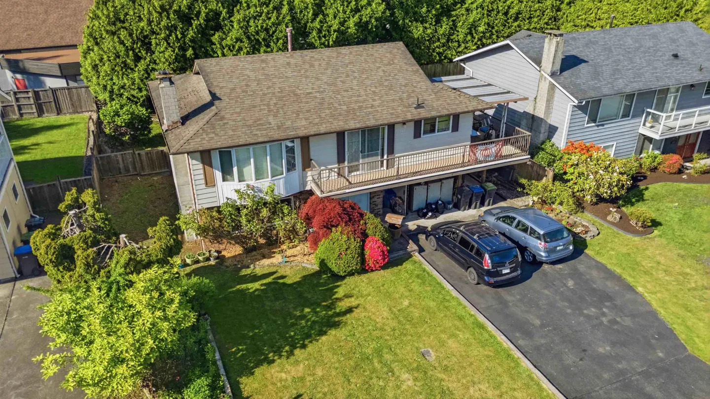 Aerial view of a two-story house with a brown roof, a balcony, and a driveway with two parked cars. The house is surrounded by green trees and grass.