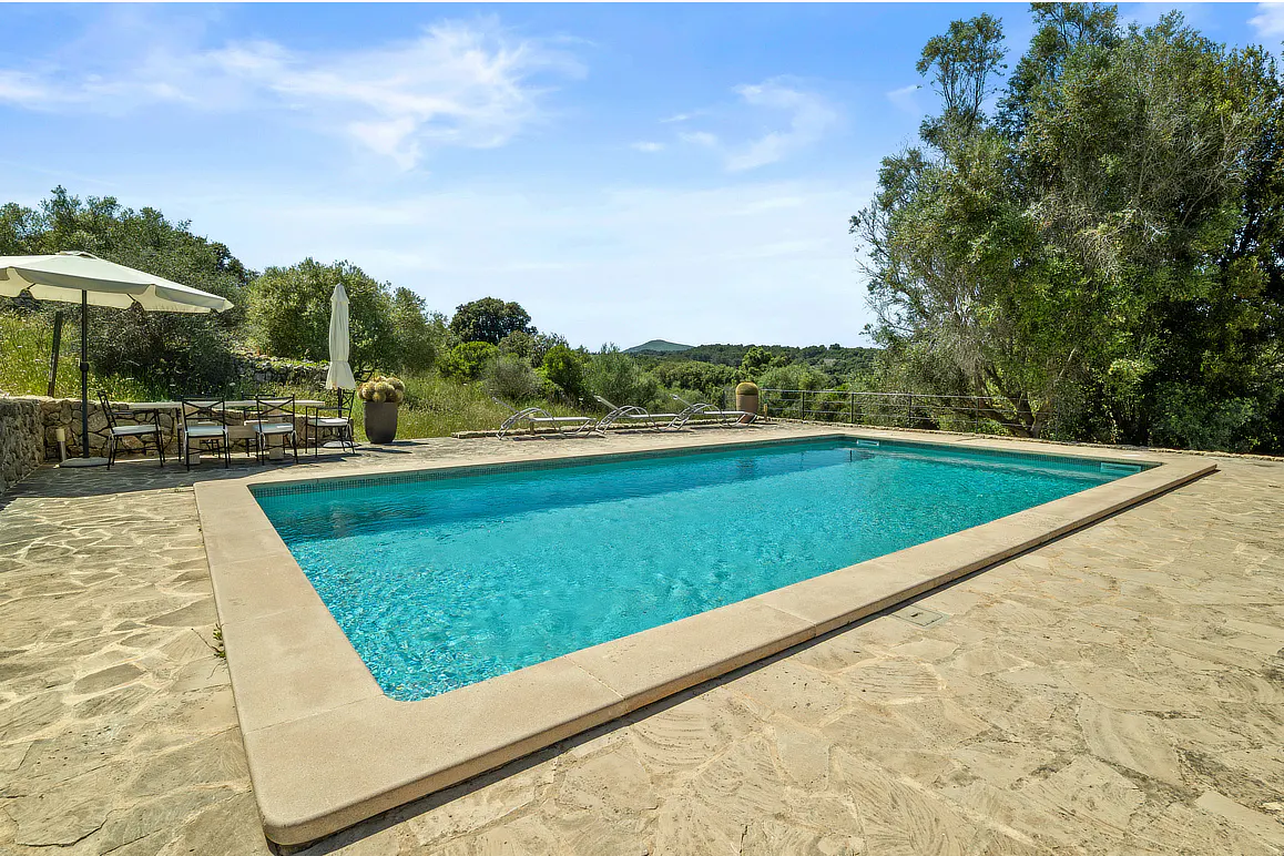 Outdoor pool with turquoise water, surrounded by stone patio, trees, and blue sky. Table and chairs under umbrella on the left.