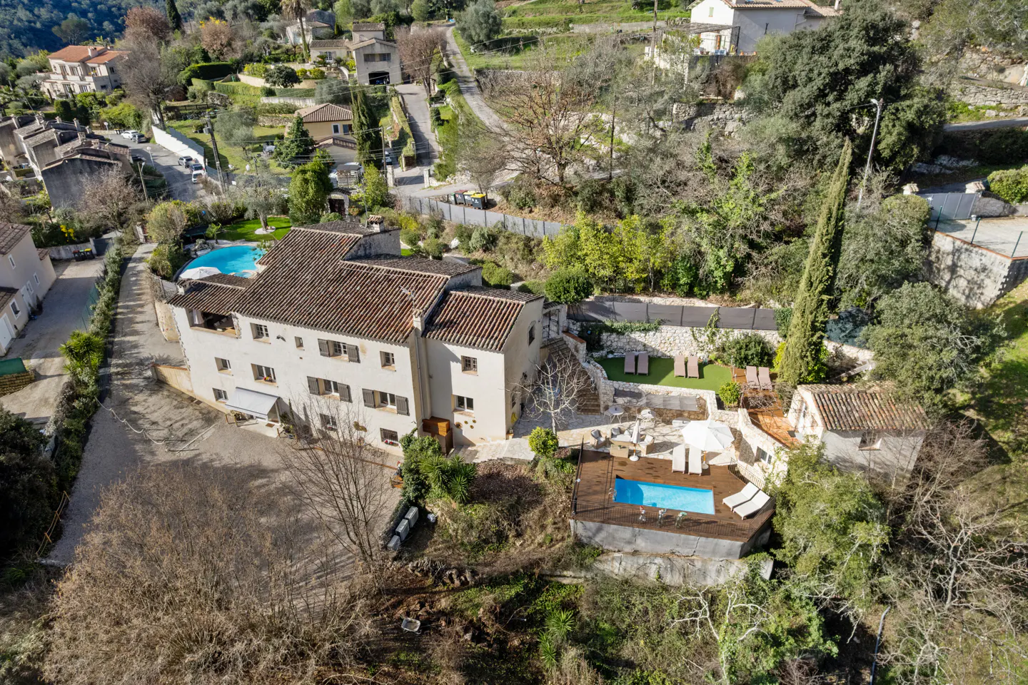 Aerial view of a beige two-story house with a brown tile roof, surrounded by trees, with two pools and lounge chairs.