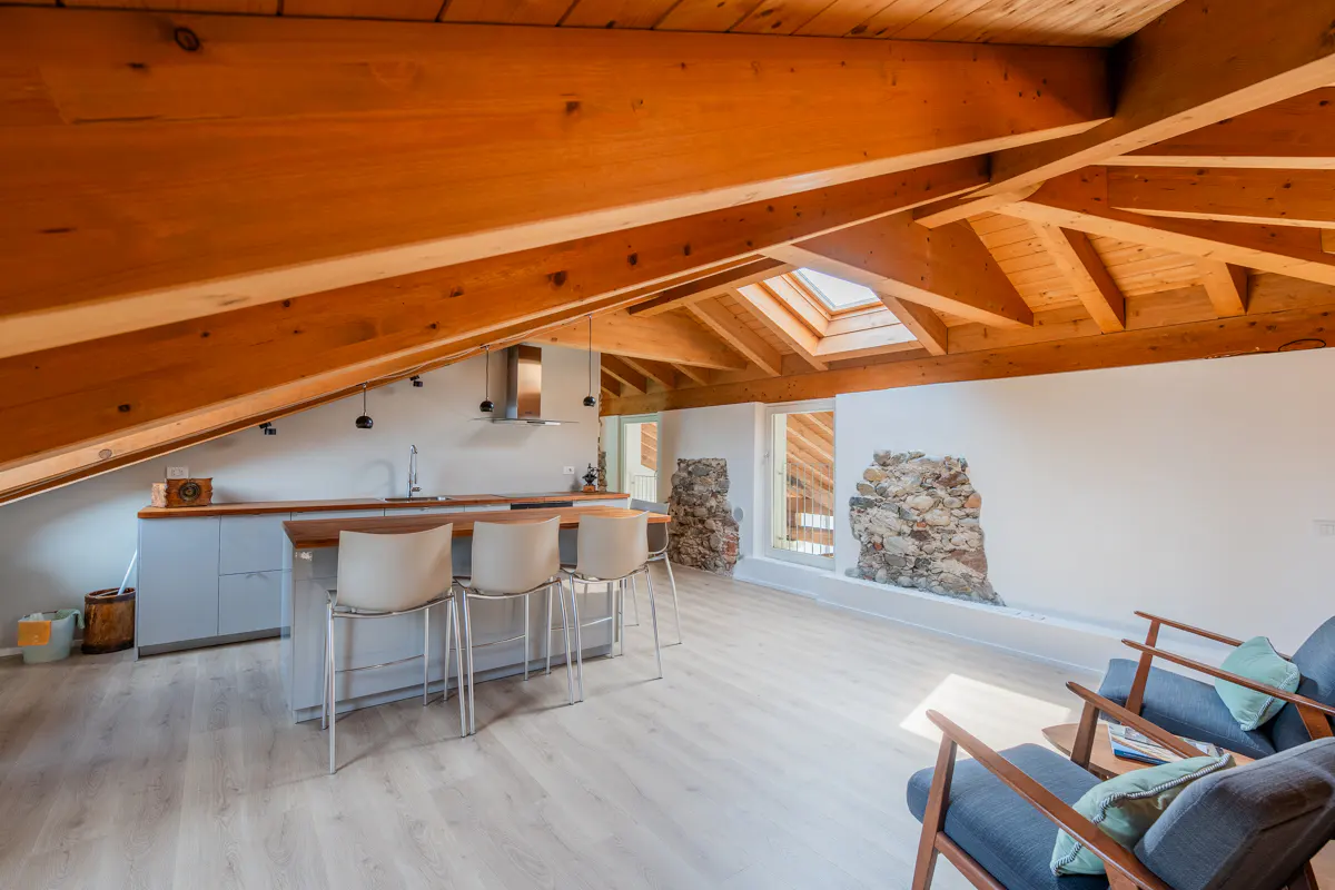 Attic apartment with exposed wood beams, skylight, kitchen island with stools, and blue armchairs.