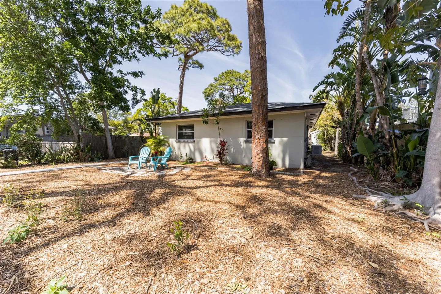 Backyard view of a one-story house with two blue chairs on a stone patio. The yard is covered in mulch and surrounded by trees.