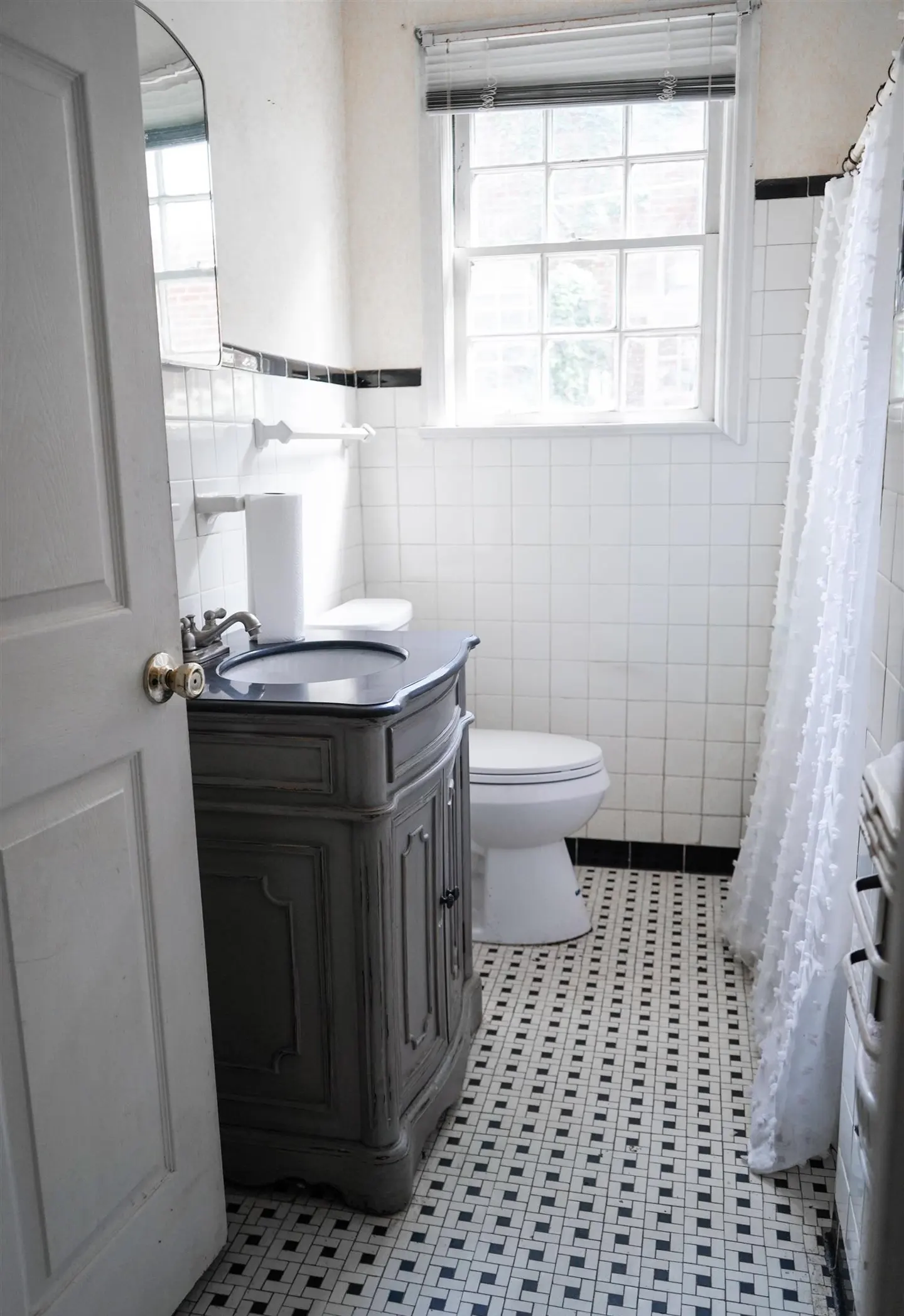 Bathroom with black and white tiled floor, white tiled walls, gray vanity, toilet, and white shower curtain.