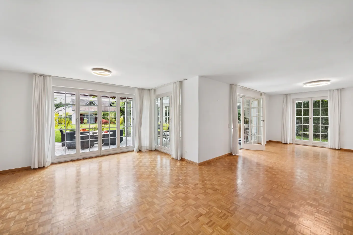 Bright, empty room with parquet floors, white walls, and multiple sets of glass doors leading to a green garden.