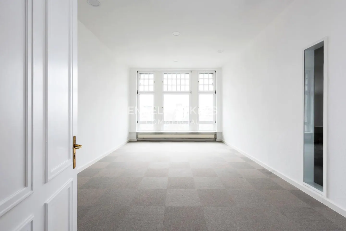 Bright, empty room with white walls, gray carpet tiles, and a large window. A white door with a gold handle is partially open.