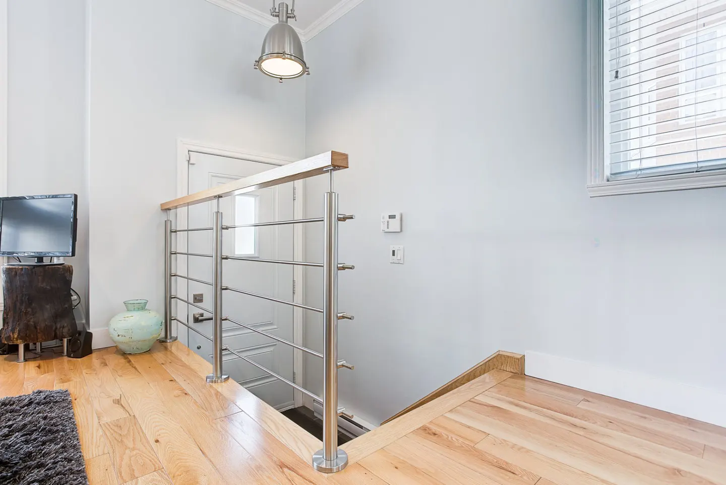 A bright landing with wood floors, a stainless steel railing, and a white door. A TV sits on a wood stump.