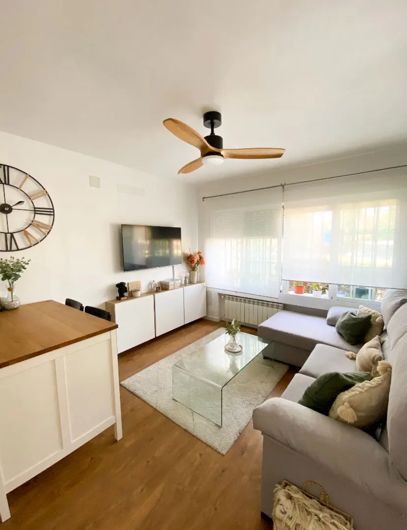 Bright living room with a grey sectional, glass coffee table, wooden dining table, and a large wall clock.