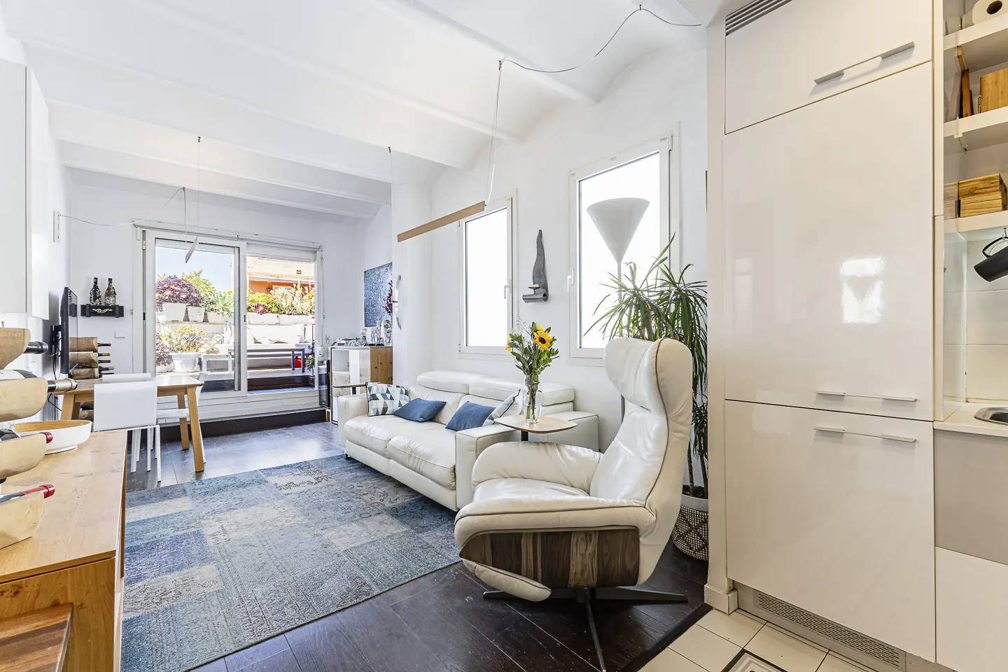Bright living room with white sofa, chair, and cabinets. A blue patterned rug sits on dark wood floors. Balcony doors open to an outdoor space.