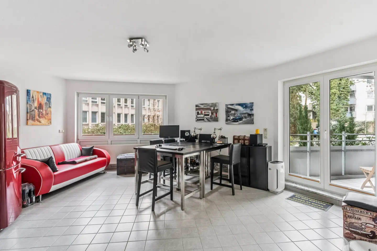 Bright living room with white walls, tiled floor, and a red retro-style sofa. A black table with chairs sits near a balcony door.