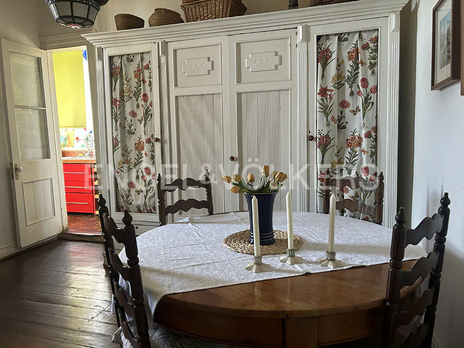 Dining room with a round wooden table, white tablecloth, vase of yellow tulips, and tall white candles. White armoire with floral curtains in the background.