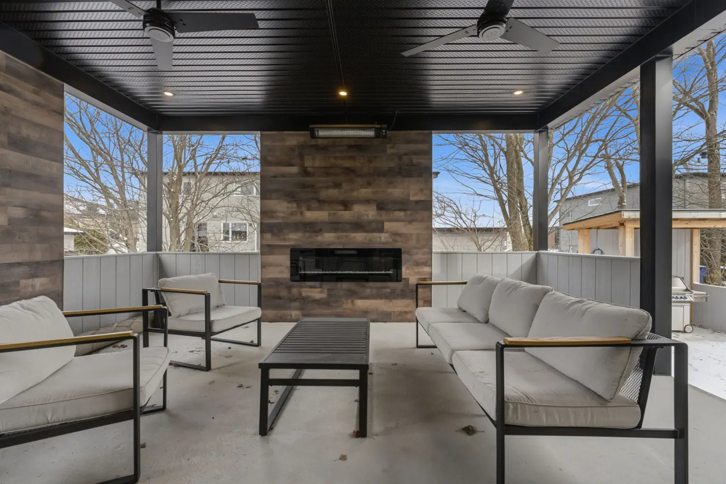 Covered patio with gray furniture, a black coffee table, and a fireplace. Two ceiling fans hang from the black ceiling. Bare trees are visible outside.