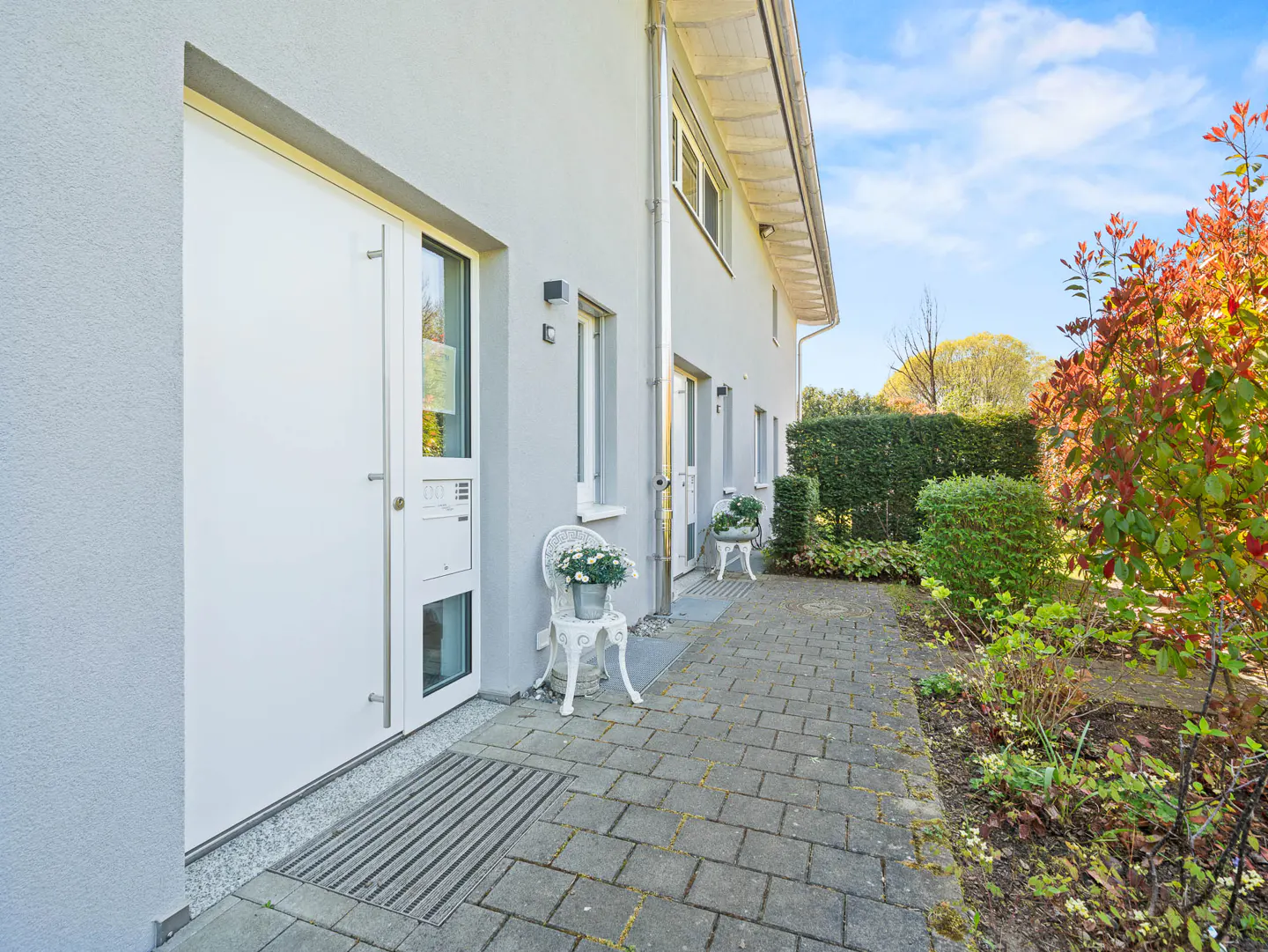 Exterior view of a modern gray house with white doors, windows, and trim. A stone walkway leads to the entrance, flanked by greenery and a white chair with flowers.
